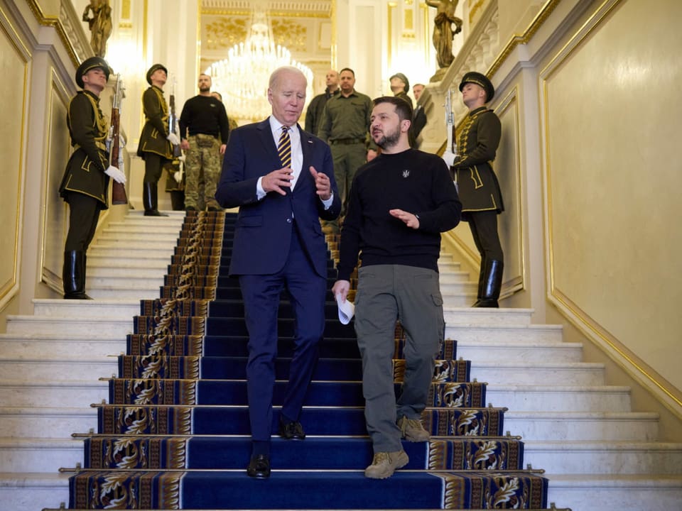 Zwei Männer gehen eine Treppe runter, auf einem dunkelblauen Teppich. Dahinter und daneben stehen uniformierte Soldaten.