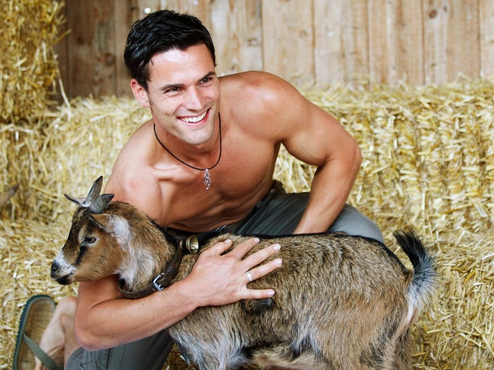 Junger Mann posiert mit nacktem Oberkörper mit einer Ziege vor Strohballen