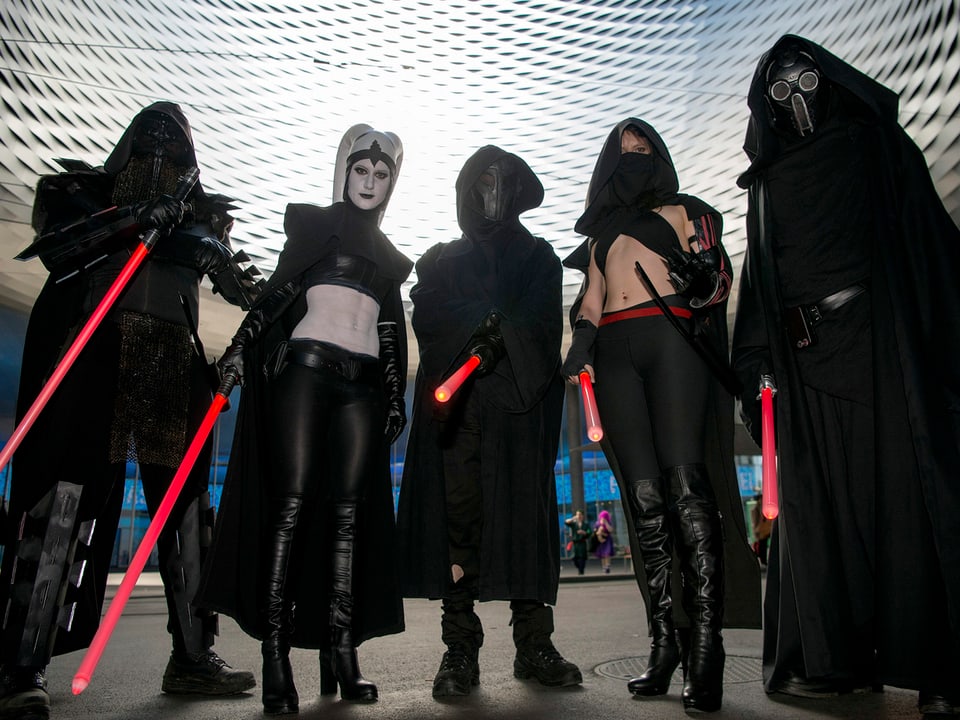 Eine Gruppe von dunkel kostümierten Männern und Frauen. Sie halten Leuchtstäbe in der Hand.