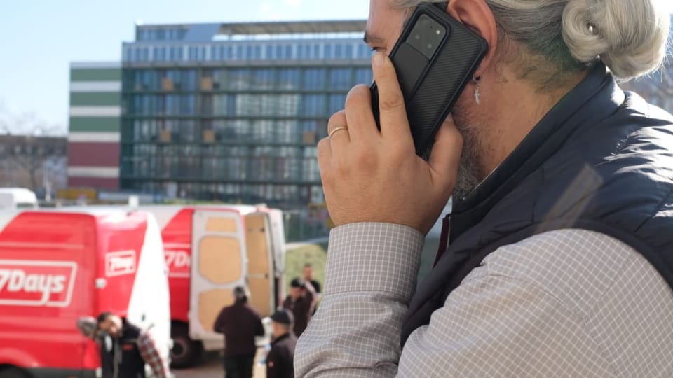 Mann mit Rossschwanz telefoniert, hinter ihm stehen mehrere Lieferwagen.