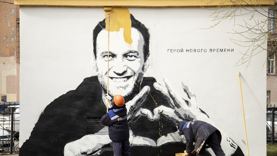 Ein Graffiti an der Wand, ein Mensch mit orangem Helm übermalt das Graffiti mit gelber Farbe.