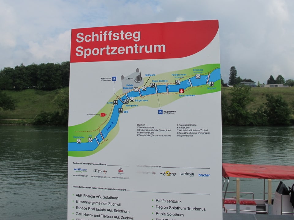 Plakat und Informationstafel der Bootsanlegestelle beim Sportzentrum Zuchwil; in einem roten Balken mit weisser Schrift steht "Schiffsteg Sportzentrum", darunter ist eine Landkarte mit dem Aareverlauf und alle anderen Anlegestellen verzeichnet.