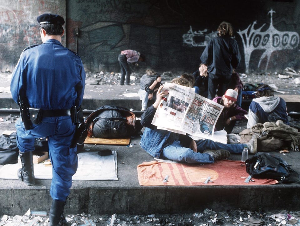 Verschiedene Randständige sitzen am Boden, lesen Zeitung und am Schlafen. Links im Bild sieht man einen Polizisten.