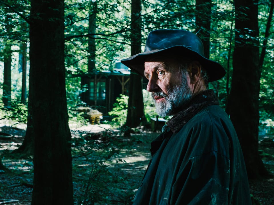 Ein älterer Mann mit Bart, Hut und dicker Jacke steht in einem Wald.