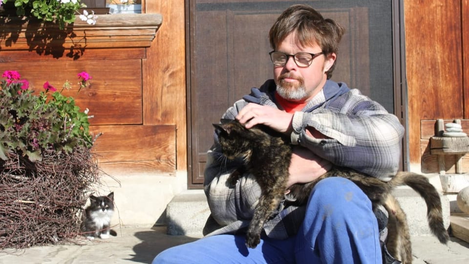Foto Mann mit Brille sitzt vor Haustür, hält dunkelbraune Katze im Arm und krault sie.