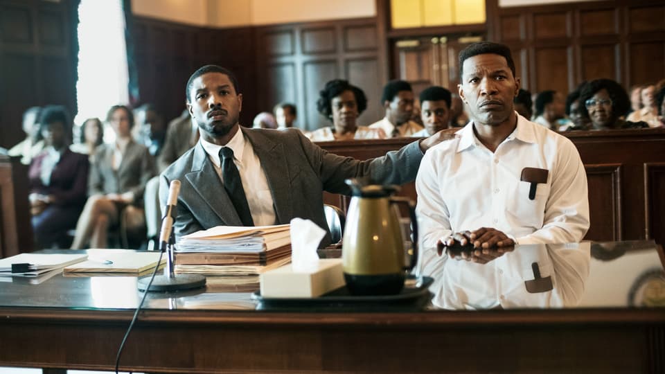 Filmszene: Zwei schwarze Männer sitzen im Gerichtssaal, einer trägt einen Anzug, der andere Sträflingskleidung.