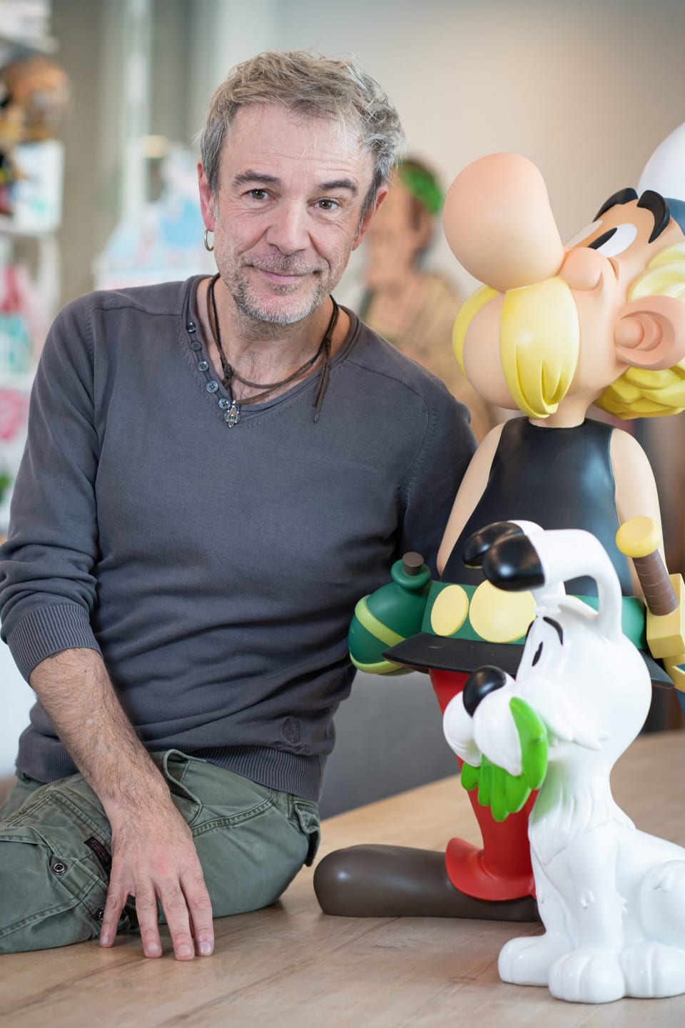 Ein Mann mit Stoppelbart und Ohrring sitzt neben den zwei Plastik-Comicfiguren Asterix und Idefix