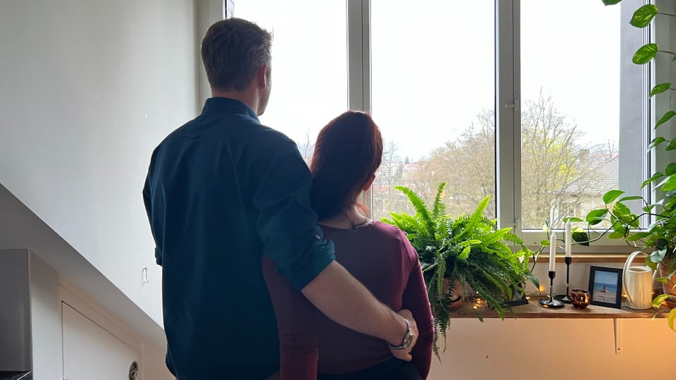 Ein Paar in ihrer Wohnung steht und blickt aus dem Fenster.