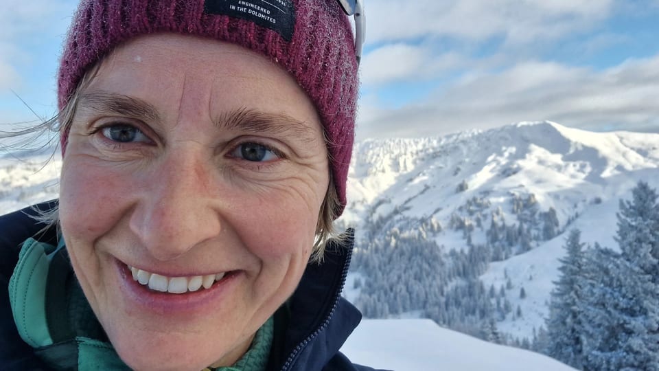 Ein Selfie von einer Frau vor einem Bergpanorama. Sie steht im Schnee. Ihr Gesicht ist ganz nah.