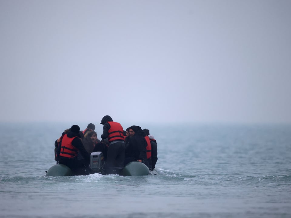 Mehrere Menschen, vereinzelt mit Rettungswesten, sitzen in einem Schlauchboot mit Motor. 