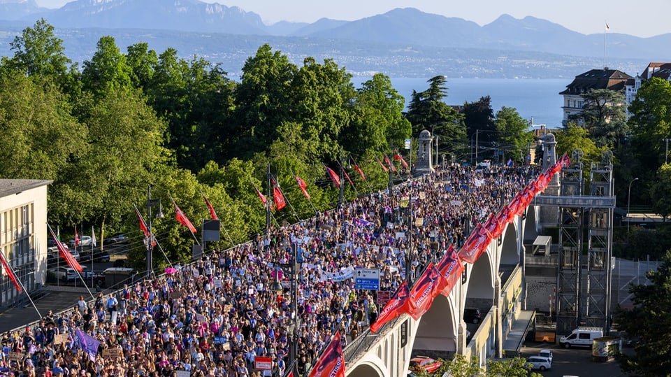 Tausende Demonstrierende ziehen über eine Brücke, im Hintergrund sind See und Bergpanorama zu sehen.