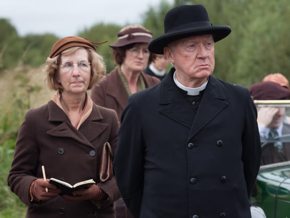 Pfarrer mit Mantel und schwarzem Hut und ernstem Blick. Neben im eine Gruppe Frauen.