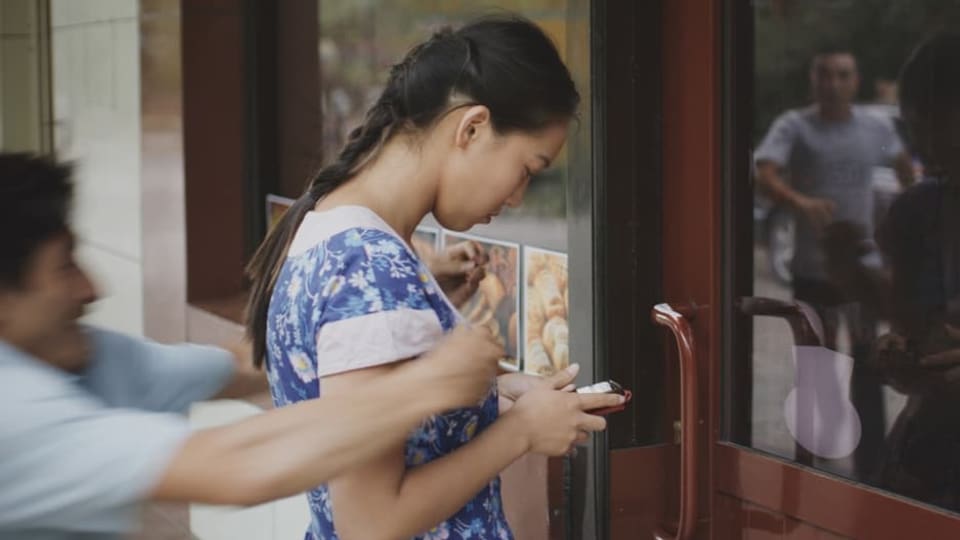 Filszene: Ein Mann greift von hinten eine junge Frau, die auf ihr Handy schaut.