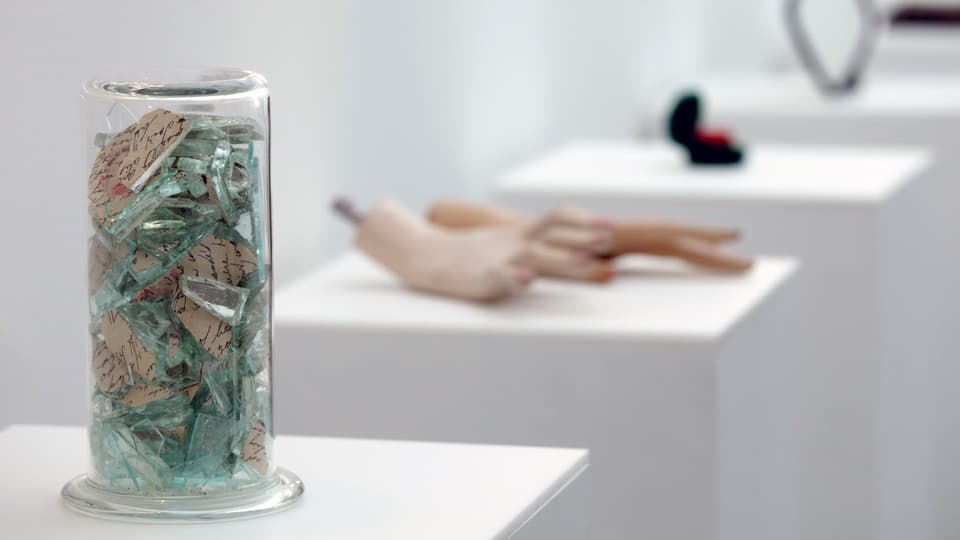 Ein Glas voller Scherben, Hände einer Puppe und weitere Objekte auf weissen Sockeln in einem Museum ausgestellt.