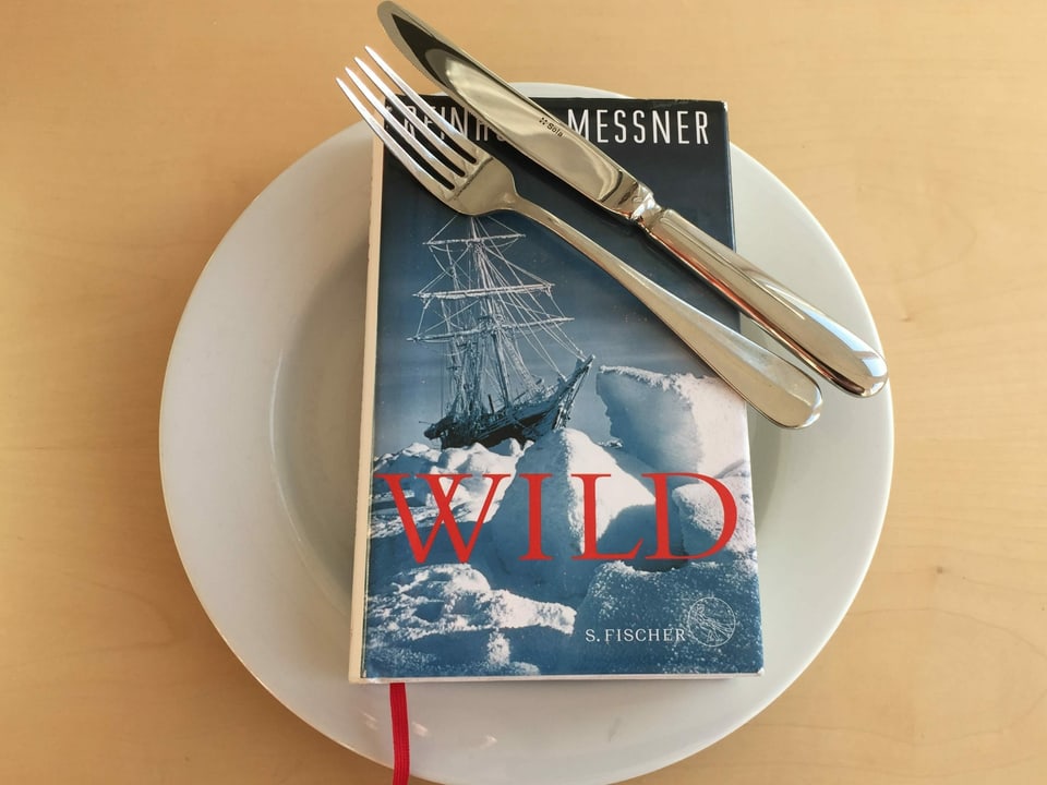 Der Abenteuerroman «Wild» von Reinhold Messner liegt auf einem weissen Teller. Messer und Gabel nebeneinander drüber.