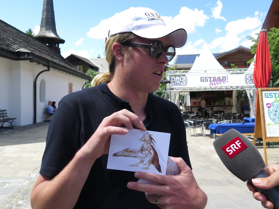 Mann mit Mütze, Brille, Postkarte mit Grafik, Mikrofon SRF in Gstaad.