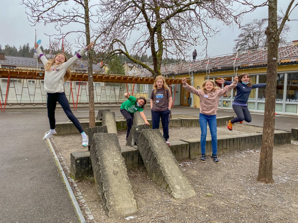 Kinder springen auf einem Schulhausplatz fröhlich von einer Mauer und strecken Arme und Beine aus. 