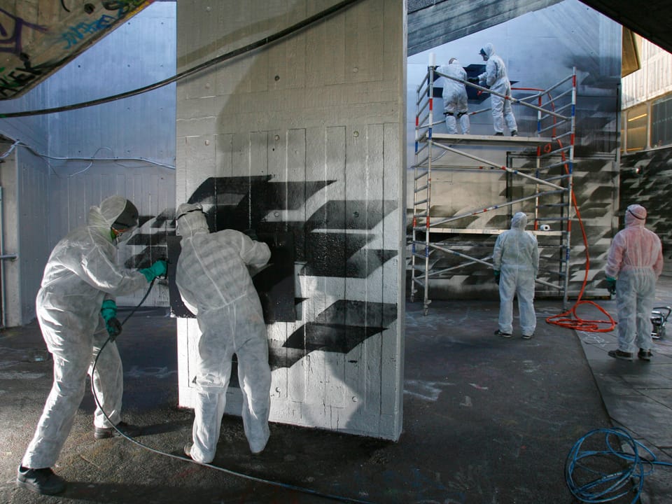 Zwei Menschen besprühen eine Wand mit schwarzen Rauten.
