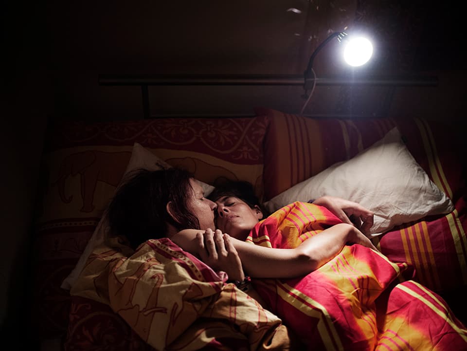 Zwei Frauen liegen im Bett unter ihren Decken und umarmen sich. 