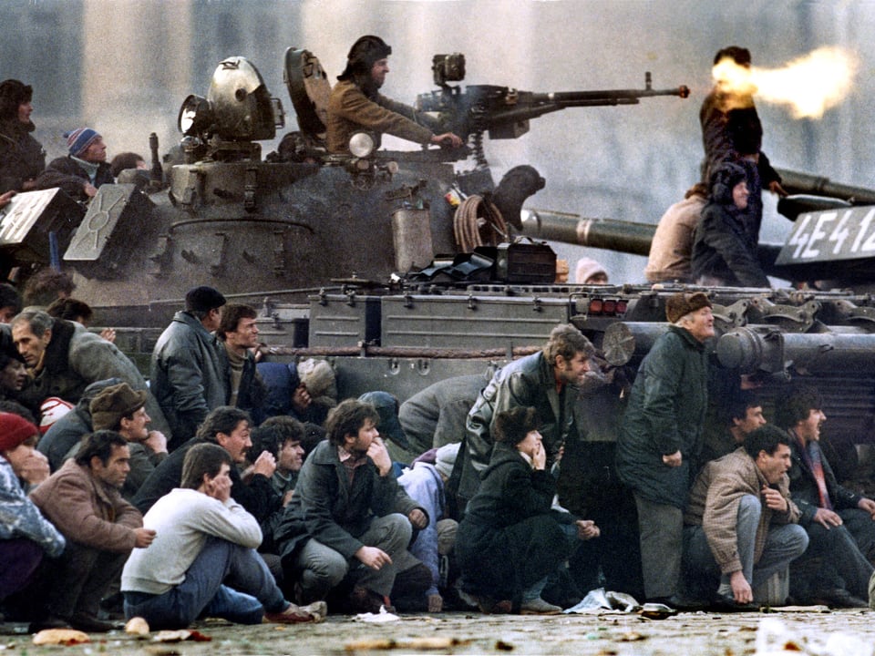 Panzer feuert, im Vordergrund sitzen Demonstranten