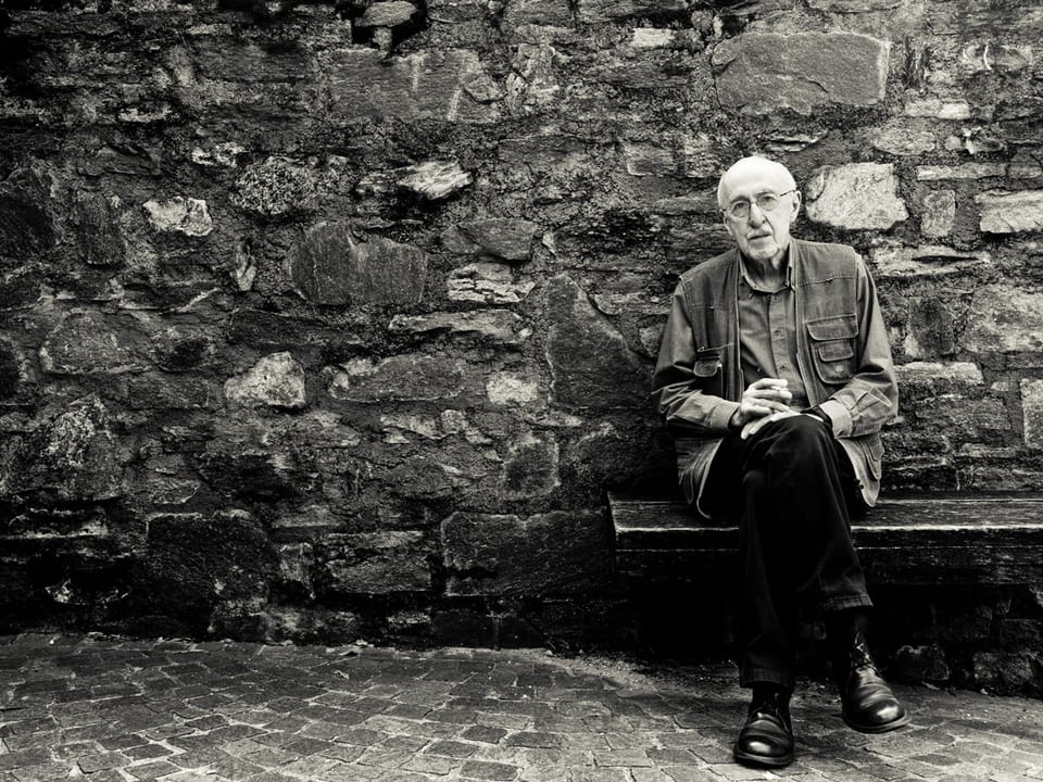 Ein älterer Herr sitzt auf einer Holzbank vor einer Steinmauer.