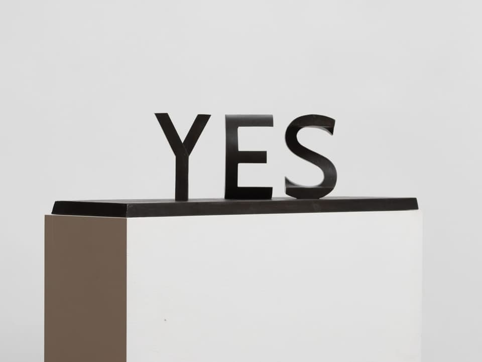 Schwarze Schriftskulptur «YES» auf einem weissen Sockel, weisser Hintergrund