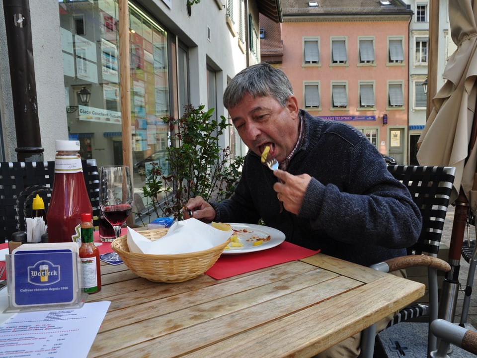 Mann schiebt Gabel mit Pommes in Mund. Restaurant aussen.