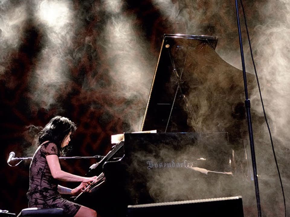 Aki Takase am Klavier spielend, umhüllt von teilweise beleuchtetem Trockeneis.