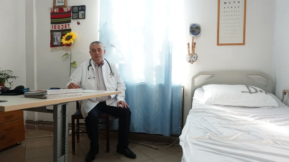 Ein Mann in weissem Kittel sitzt an einem Pult daneben ein Spitalbett