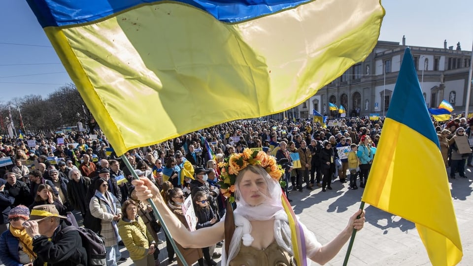 Eine Frau schwenkt an einer Demo zwei Ukraine-Flaggen, dahinter sind viele Menschen.