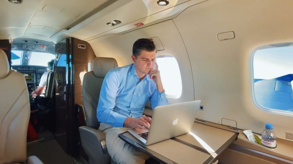 mittelalter Mann in blauem Hemd sitzt in Privatjet, weisse Ledersessel, ein silbernes Macbook vor sich.
