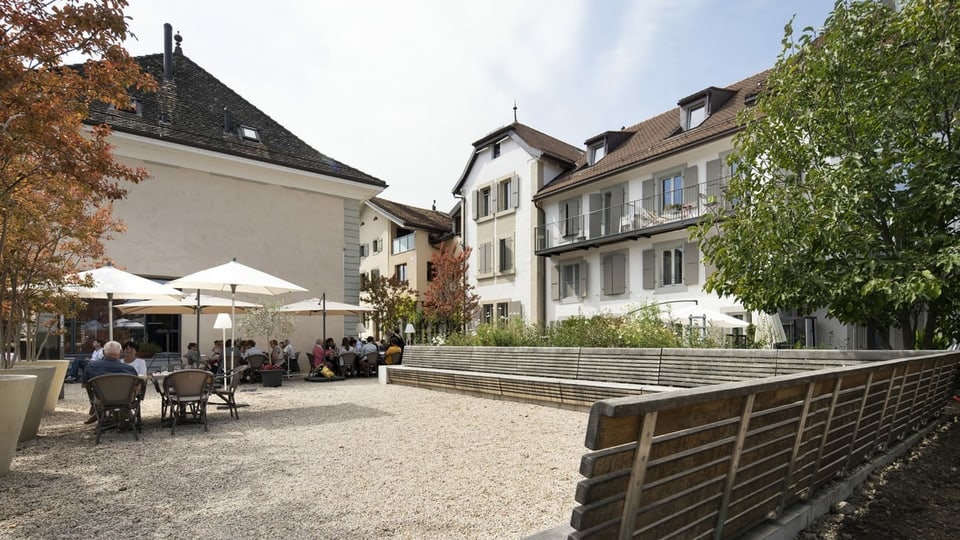 Blick auf einen Dorfplatz mit Tischen und Stühlen und historischen Gebäuden im Hintergrund.