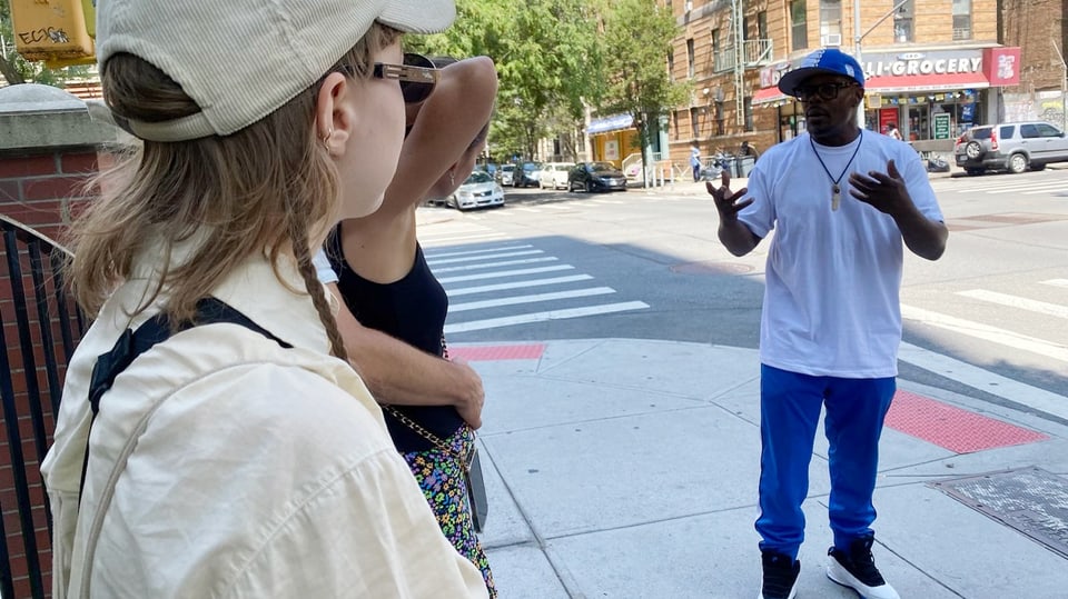 Schwarzer Mann in mittlerem Alter mit weissem T-Shirt und Cap, Hände erklärend in der Luft, auf dem Gehsteig einer Stadt
