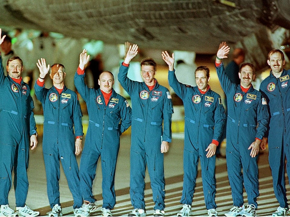 Sieben Astronauten winken nebeneinanderstehend in die Kamera.