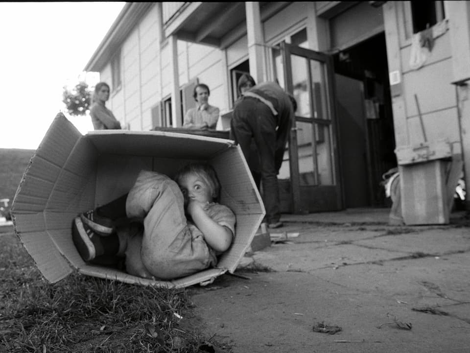 Ein Kind sitzt in einer kartonschachtel. Im Hintergrund ist der Eingang zu den Sozialwohnungen. 