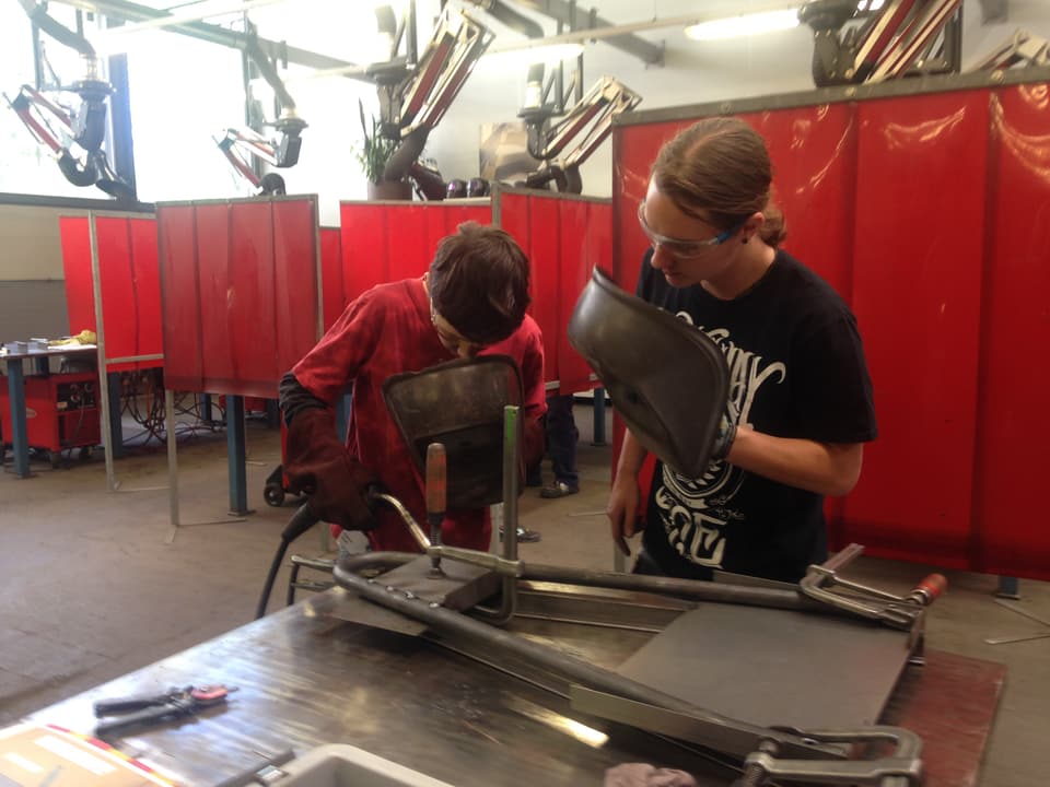 Ein Junge und ein Lehrling arbeiten an einem Metallstück, in der Hand halten sie einen Sichtschutz. 