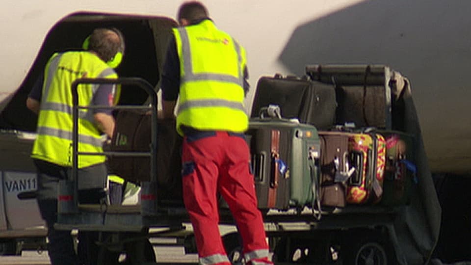 Zwei Gepäckarbeiter mit gelber Warnweste beladen von Hand ein Flugzeug mit Gepäck, das auf einem Wagen liegt