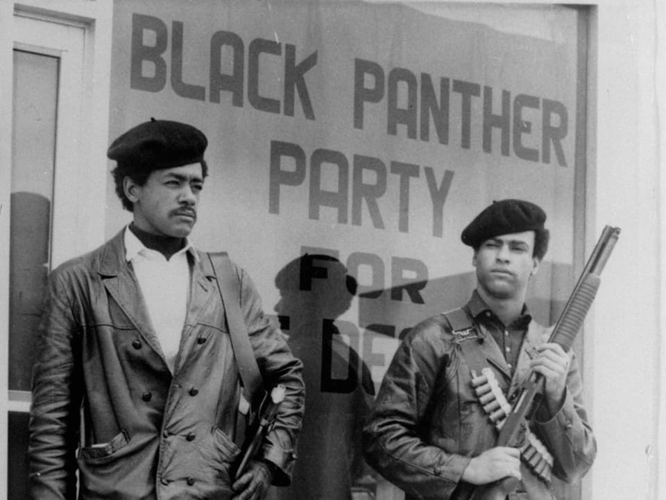 Zwei schwarte bewaffnete Männer stehen vor einer Glasscheibe mit dem Schriftzug «Black Panther Party»