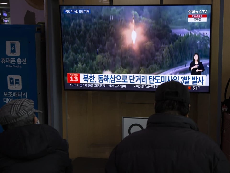 Ein TV-Bildschirm zeigt ein Bild einer startenden Rakete. Vor dem TV sitzen Menschen.