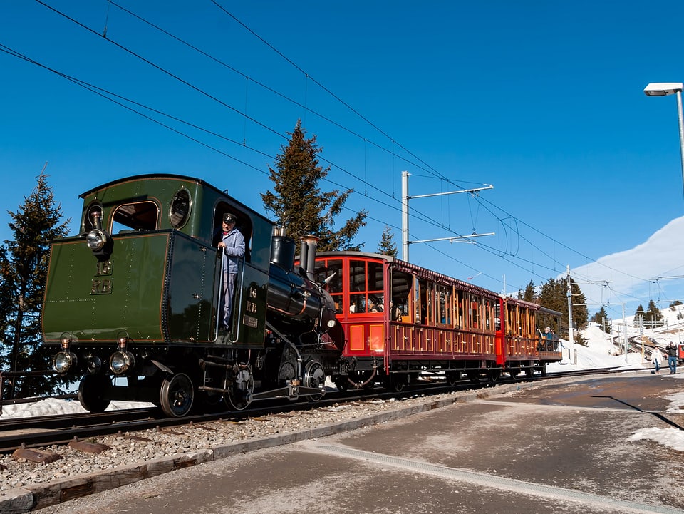 In schneebedeckter landschaft zieht eine grüne alte Dampflok einen knallrot angestrichenen alten Bahnwagen.