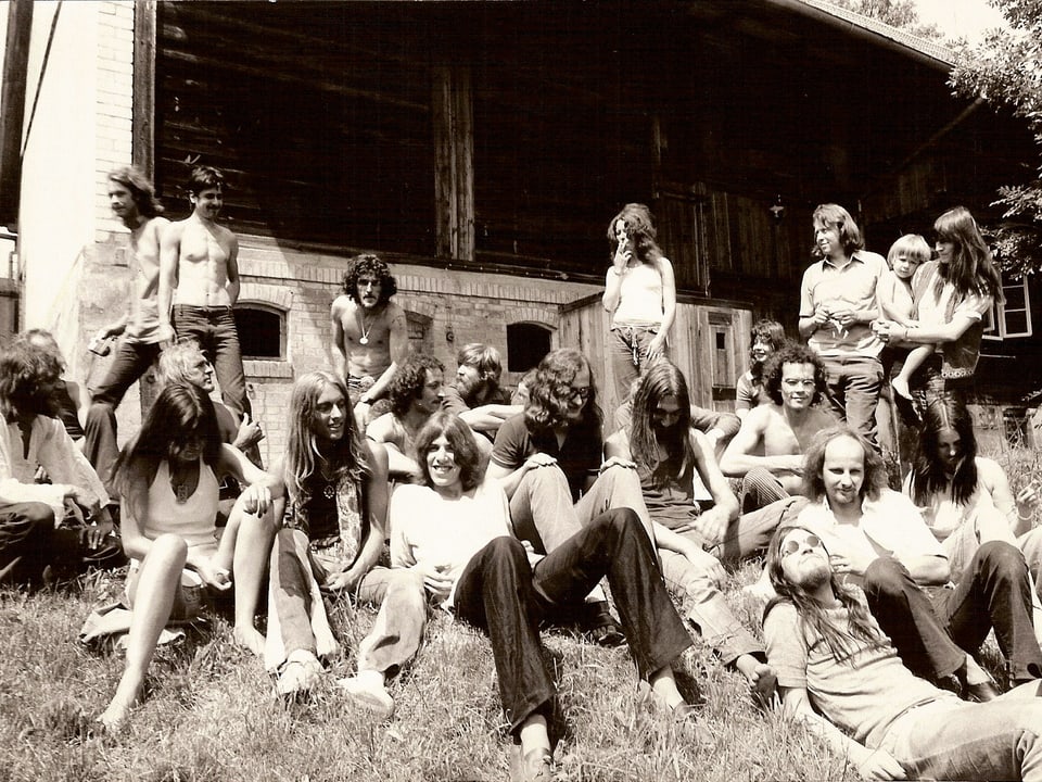 Eine grössere Gruppe von Menschen mit langen Haaren auf einer Wiese vor einem Holzhaus.