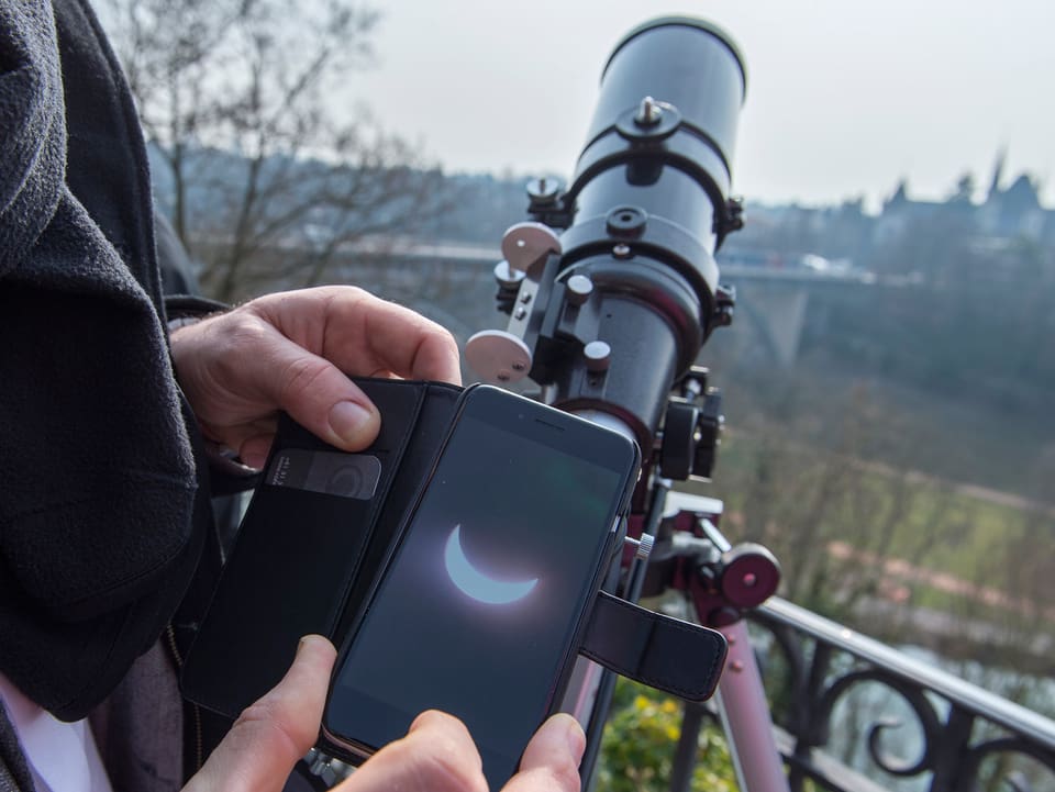 Zu sehen ist ein Fernglas mit einer Handycamera, welche die Sonnenfinsternis einfängt.