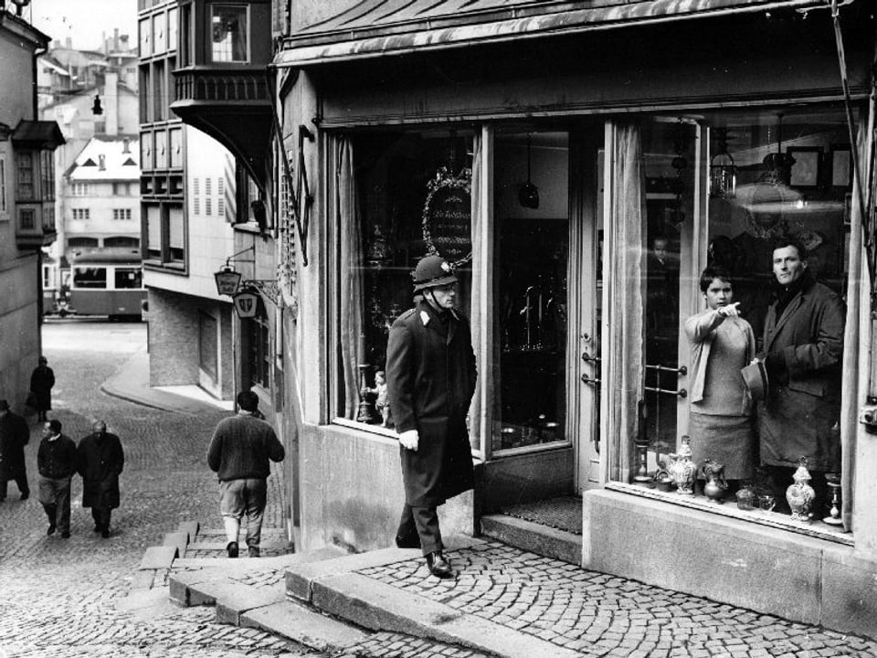 Eine Gasse im Züricher Niederdorf. Ein Polizist in Uniform geht auf einen Laden zu. Im Schaufenster stehen eine junge Frau, die mit dem Finger auf etwas zeigt, und ein Mann im Mantel.