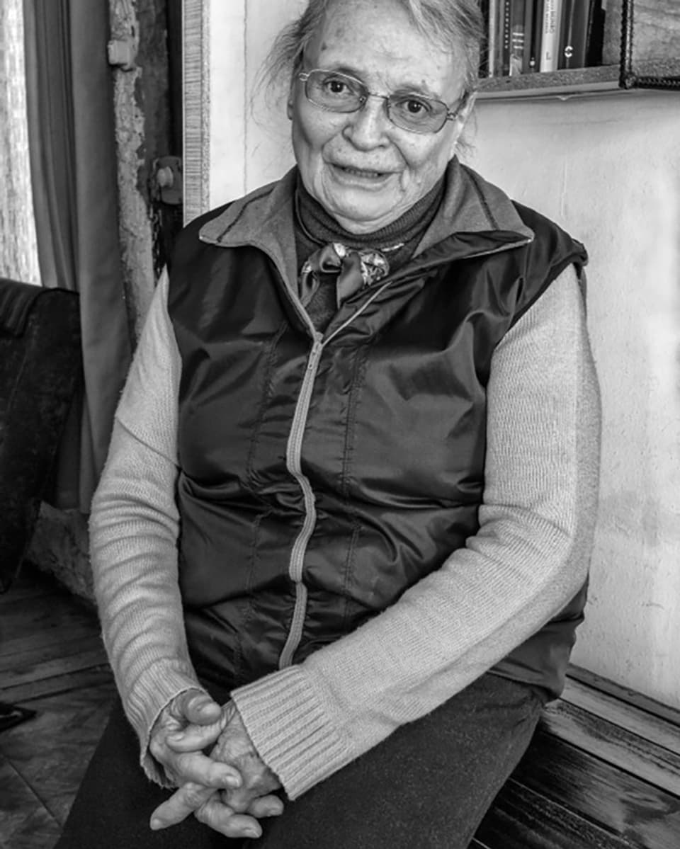 Alte Frau mit Brille sitzt auf einer Bank, die Hände verschränkt. Sie trägt Rollkragenpullover, Halstuch und Weste.