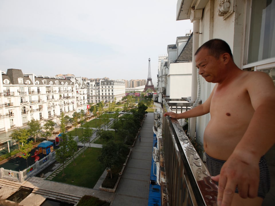 Mann mit nacktem Oberkörper steht auf Balkon seiner Wohnung und schaut in den Park hinunter.
