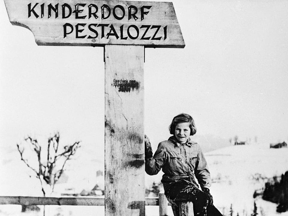Ein Mädchen sitzt neben einem Schild mit der Aufschrift: «Kinderdorf Pestalozzi»