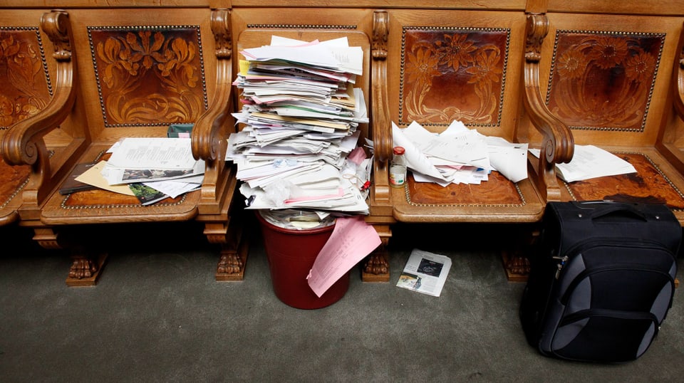 Ein Papierstapel und weitere Papiere, auf mehrere Sitze aus Holz im Parlament verteilt.