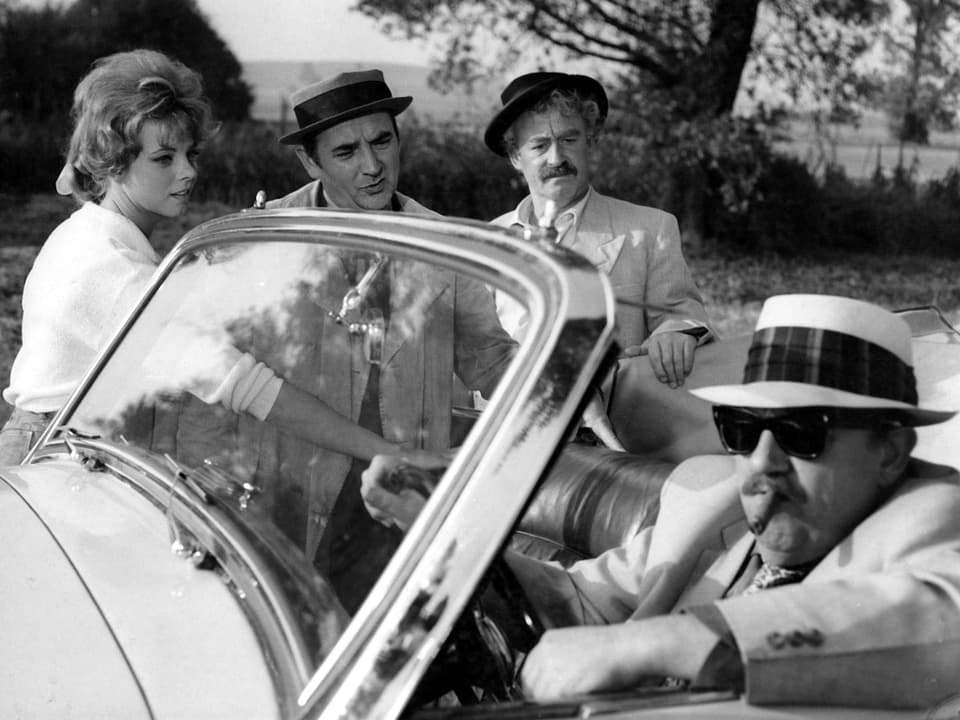 Ein Mann mit Zigarre im Mund und Hut auf dem Kopf sitzt in einem Cabriolet. zwei Männer und eine Frau stehen neben dem Auto.
