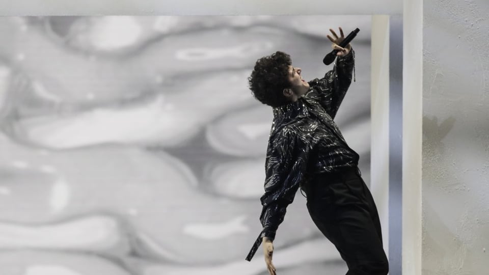 Person in schwarzem Outfit hält Mikrofon und gestikuliert beim Singen vor abstraktem Hintergrund.