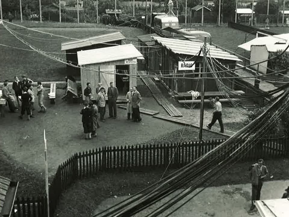 Die Schwarz-Weiss-Fotografie zeigt Menschen und kleine Holzbaracken auf einer Wiese.
